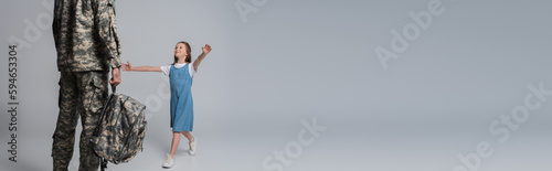 happy girl welcoming father in military uniform with backpack during homecoming on grey background, banner.