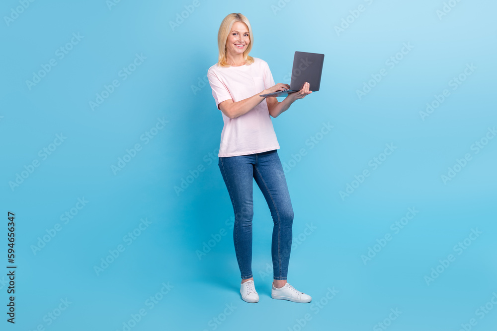 Full size photo of positive nice senior woman white t-shirt jeans sneakers writing letter on laptop isolated on blue color background Full size photo of positive nice senior woman white t-shirt jeans sneakers writing letter on laptop isolated on blue color background