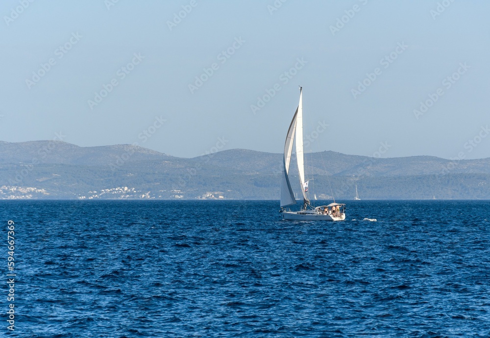 White sailboat glides through a vast ocean with a picturesque shoreline in the background.