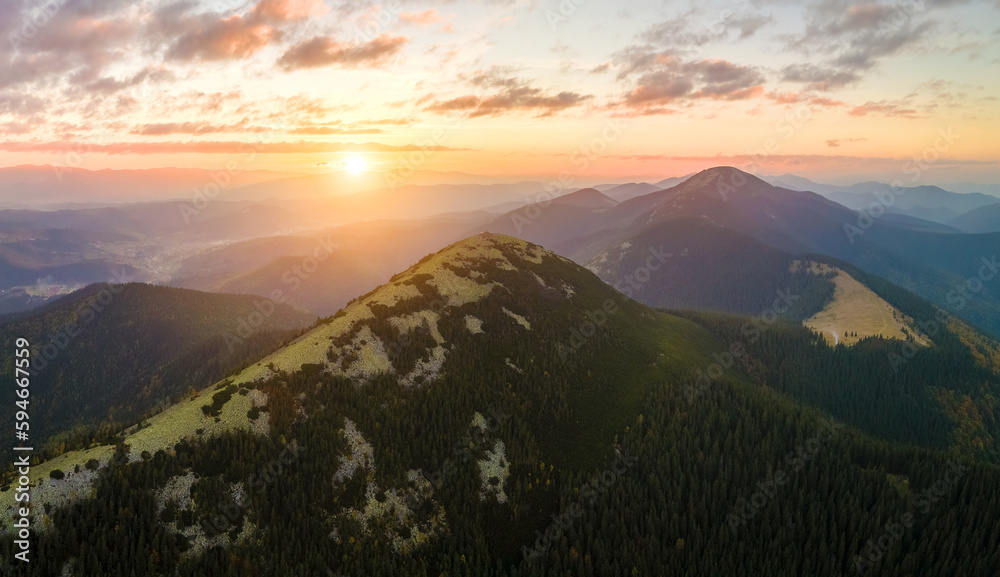 Aerial view of amazing scenery with foggy dark mountain peak covered with forest pine trees at autumn sunrise. Beautiful wild woodland with shining rays of light at dawn