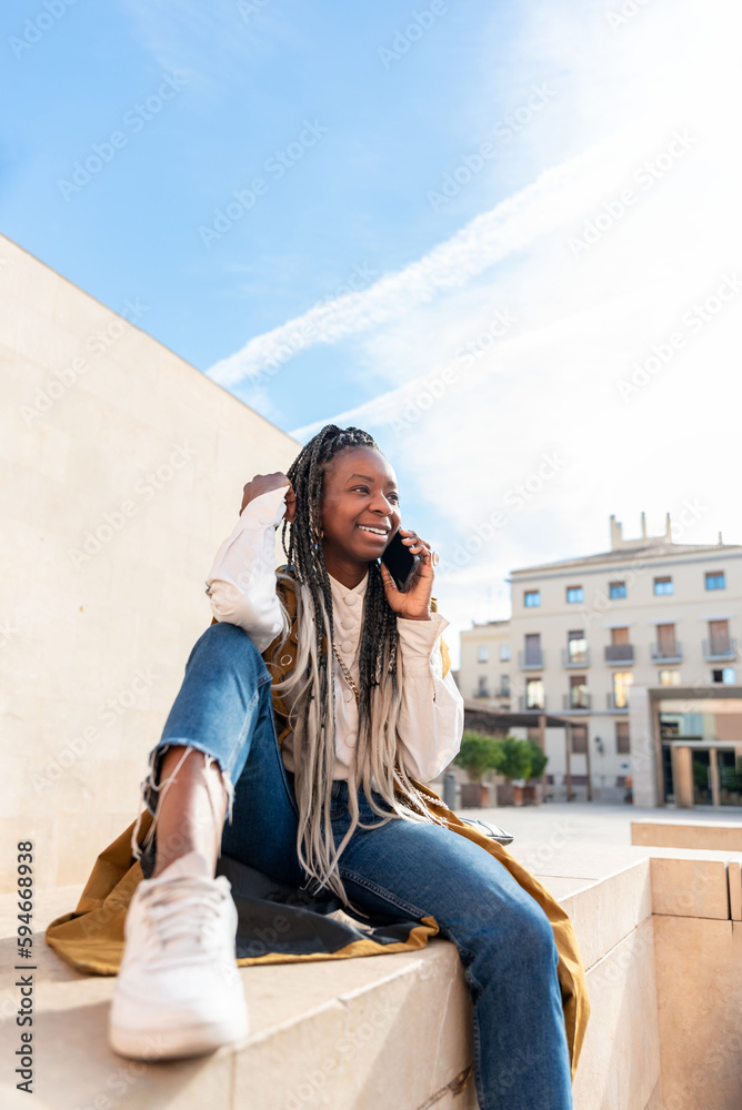 a smiling african american woman with braids sitting on a wall outdoor against a clear blue sky and talks by mobile phone