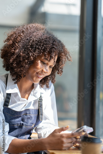 Attractive african american young business owner or barista in apron sits relaxing with mobile phone browsing apps in front of coffee shop counter.