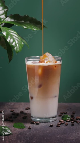 Pouring caramel sauce into a glass with iced coffee on dark teal background with coffee leaves