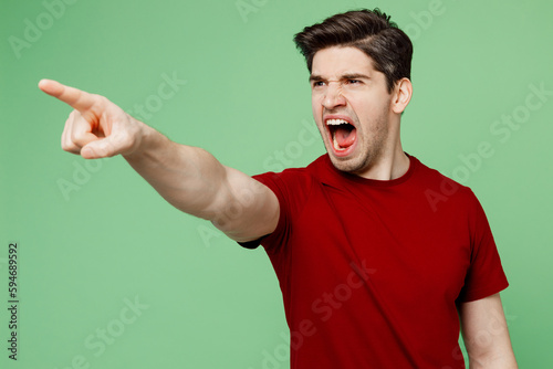 Young dissatisfied mad sad brunet caucasian man he wears red t-shirt casual clothes point index finger aside scream isolated on plain pastel light green background studio portrait. Lifestyle concept.