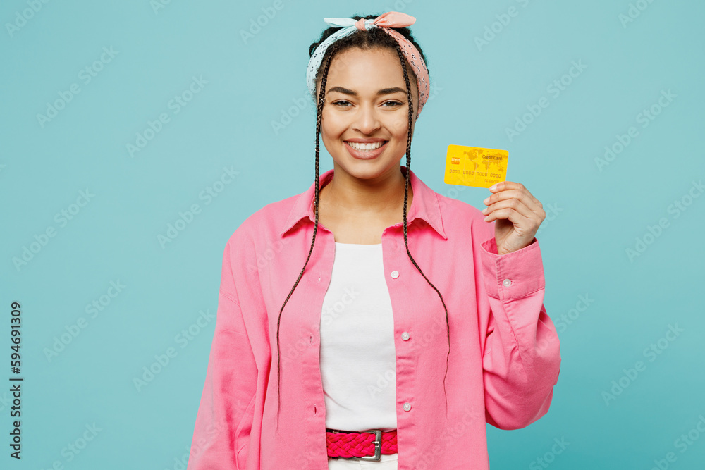 Young smiling woman of African American ethnicity wear pink shirt white t-shirt headscarf hold in hand mock up of credit bank card isolated on plain pastel light blue cyan background studio portrait. Young smiling woman of African American ethnicity wear pink shirt white t-shirt headscarf hold in hand mock up of credit bank card isolated on plain pastel light blue cyan background studio portrait.