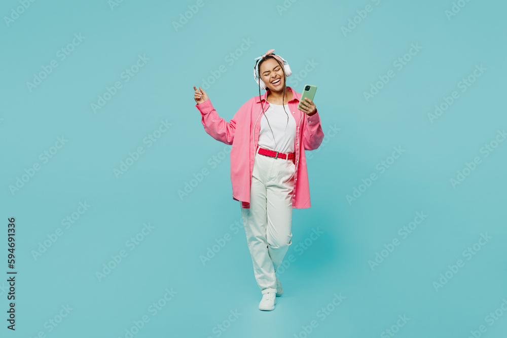 Full body young woman of African American ethnicity wear pink shirt white t-shirt headscarf headphones listen to music use mobile cell phone isolated on plain pastel light blue cyan background studio.