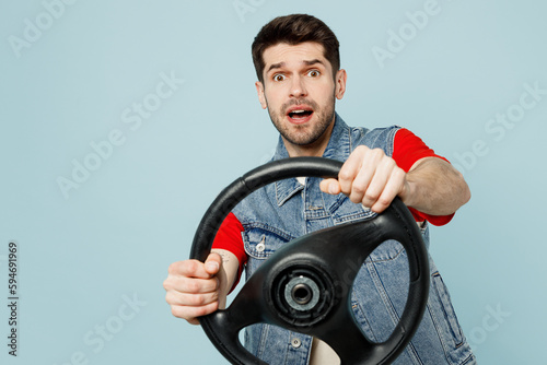 Young shocked scared man he wear denim vest red t-shirt casual clothes hold steering wheel driving car look aside on area isolated on plain pastel light blue cyan background studio. Lifestyle concept.