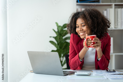Elegant african american businesswoman in formal attire sitting with hot cup of coffee during break to relax and drinking anti-drowsy coffee while working on laptop recording meeting in office.