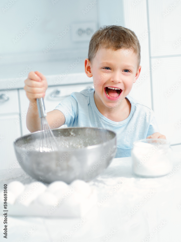 Child cooks food. Caucasian boy sits at table in kitchen and kneads dough in cup with hand whisk. Cheerful and positive kid cooks pie and laughs. Young cook or chef. Portrait of child.. Child cooks food. Caucasian boy sits at table in kitchen and kneads dough in cup with hand whisk. Cheerful and positive kid cooks pie and laughs. Young cook or chef. Portrait of child..