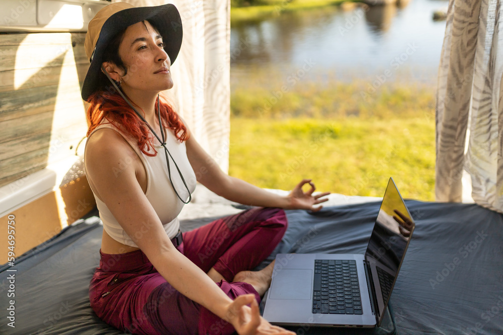 Red-haired woman teleworking typing with laptop in camper van in middle of nature near lake, connected to internet, rested from work doing meditation.