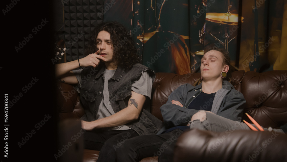 Musical rock band sits on couch. Stylish male musicians, artists, performers listen to ready recorded songs for new album in professional sound and music recording studio. Concept of music production.