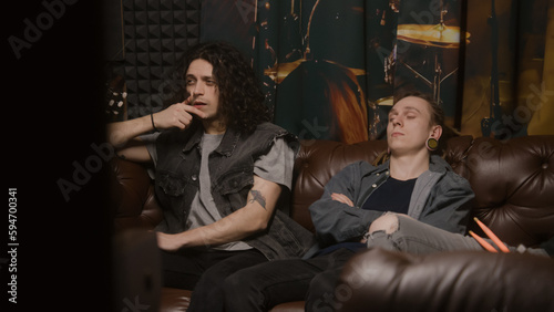Musical rock band sits on couch. Stylish male musicians, artists, performers listen to ready recorded songs for new album in professional sound and music recording studio. Concept of music production.