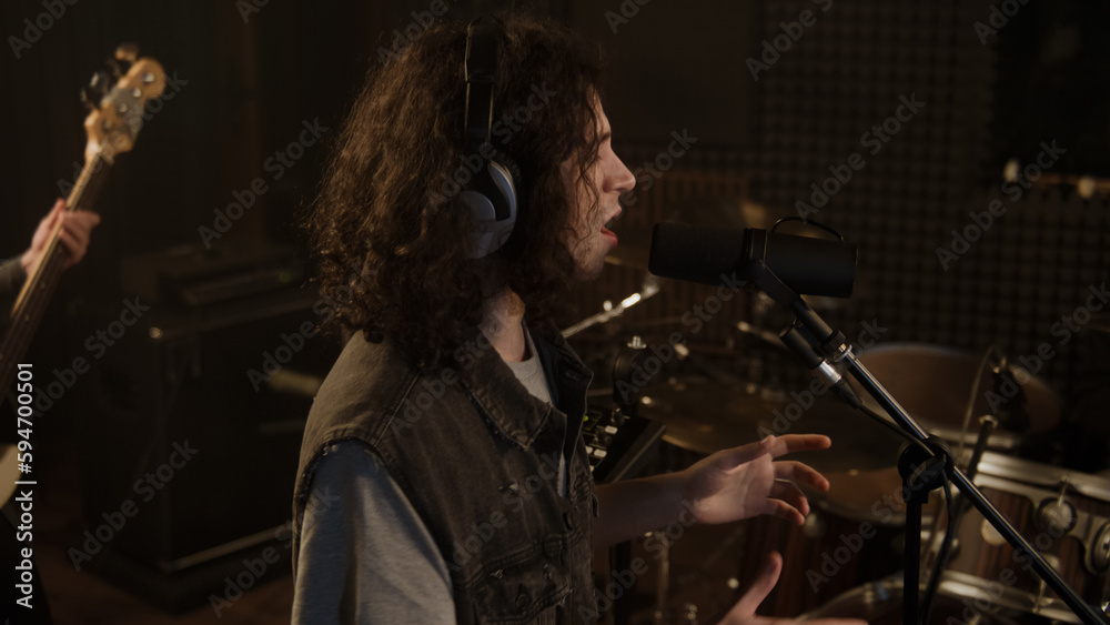Vocalist in professional headphones sings composition into microphone in soundproof room. Musicians play on guitars and drum set. Cool rock band in sound recording studio. Concept of music production.