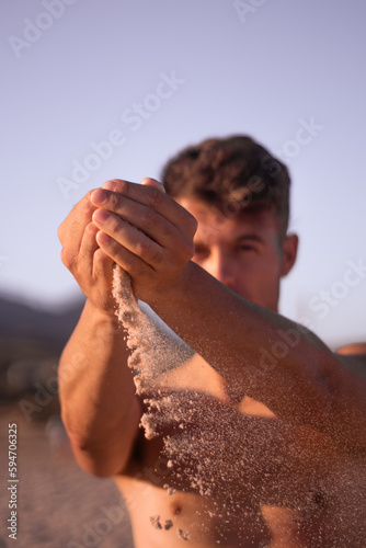 a young man with slanted eyes and white skin gently lets the sand of the beach fall between his fingers on a vacation day.
