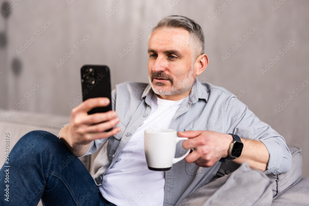 Man drinking coffee and using mobile phone at home. Adult caucasian male enjoying morning routine at living room sofa Man drinking coffee and using mobile phone at home. Adult caucasian male enjoying morning routine at living room sofa