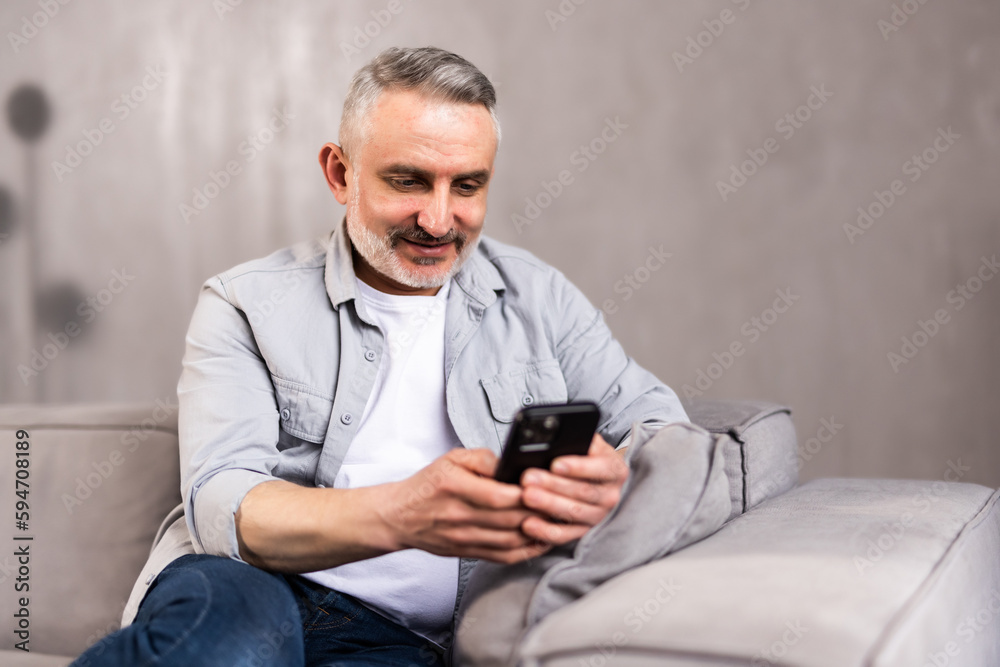 Smiling mature grey-haired man sitting on couch and using mobile phone at home, copy space. Cheerful middle-aged man with smartphone, chatting with his family or using mobile application Smiling mature grey-haired man sitting on couch and using mobile phone at home, copy space. Cheerful middle-aged man with smartphone, chatting with his family or using mobile application