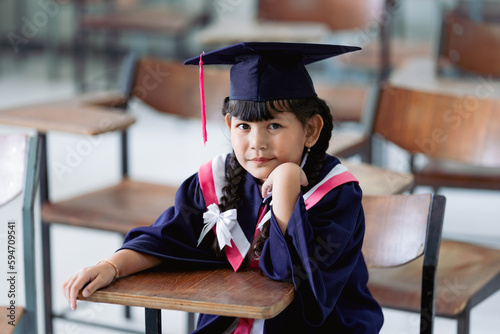 A cheerful kid graduate wearing an academic gown and graduation cap celebrates her graduation in the classroom. Education stock photo