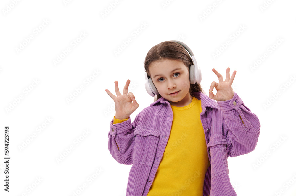 Elementary age child, lovely little school girl in casual clothes, enjoys listening music on wireless headphones, gestures with her hands, shows OK sign, smiles at camera, isolated white background Elementary age child, lovely little school girl in casual clothes, enjoys listening music on wireless headphones, gestures with her hands, shows OK sign, smiles at camera, isolated white background