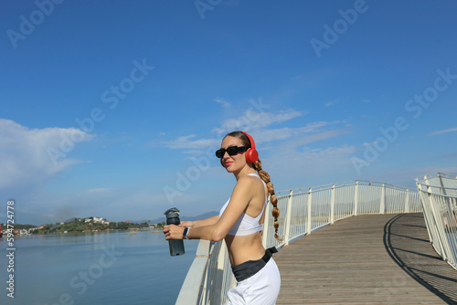 Fit woman in her 30s standing on the bridge with the water bottle, wearing white sportswear and red headphones. Background, copy space for text, close up. © Evrymmnt