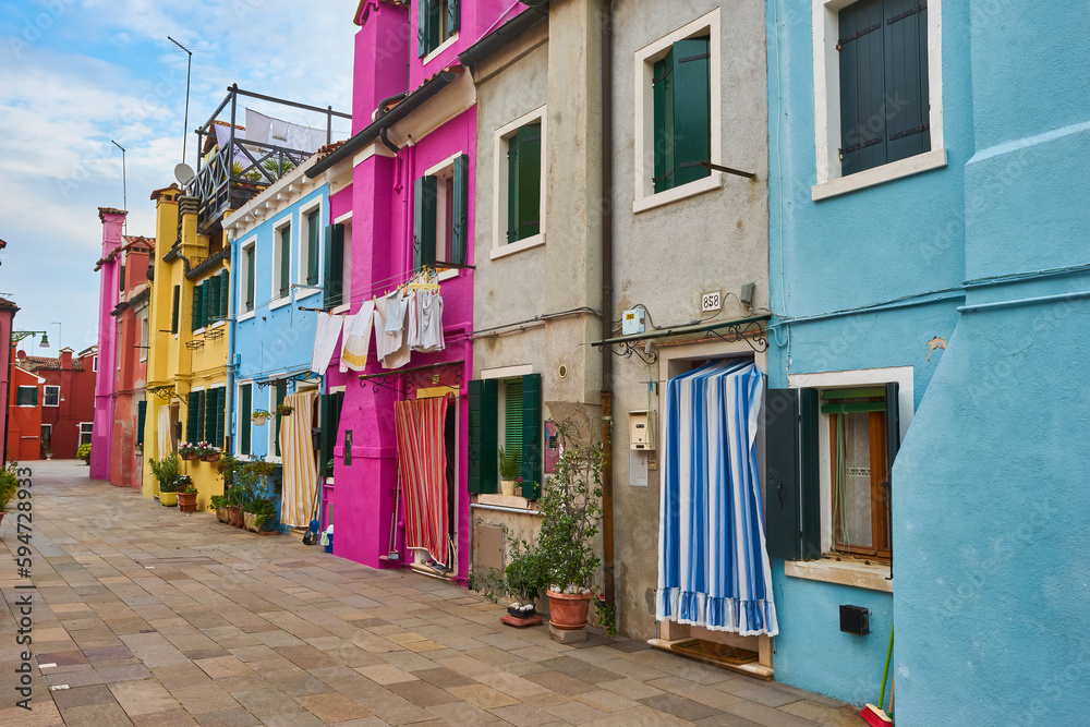 Burano island narrow cobblestone street between colorful houses buildings with multicolored bright walls, blue sky sunny summer day, Venice Province, Veneto Region, Northern Italy.