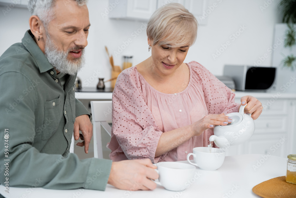 joyful woman pouring tea near bearded husband smiling in kitchen. joyful woman pouring tea near bearded husband smiling in kitchen.