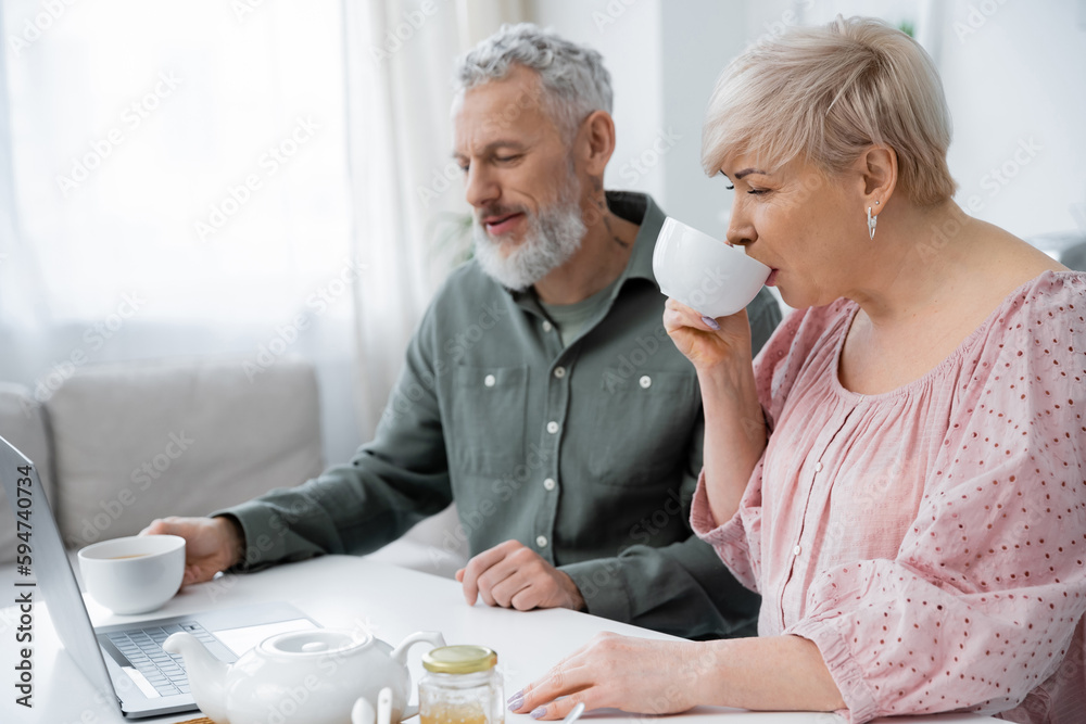 Obraz premium positive middle aged couple looking at laptop while enjoying morning tea in kitchen.