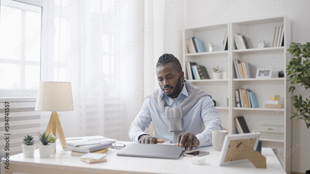 An African American man is sitting at his remote workspace with his laptop at home An African American man is sitting at his remote workspace with his laptop at home