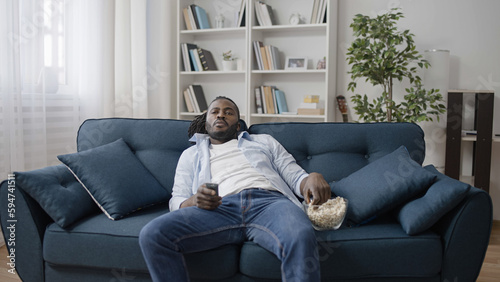Fényképezés Carefree African American man eating popcorn, lying on sofa and watching TV