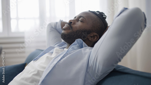 Tired African American man sitting back and relaxing on sofa at home after work day