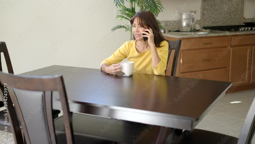 Attractive cheerful woman relaxing talking on mobile cell phone sitting at dining table drinking coffee Attractive cheerful woman relaxing talking on mobile cell phone sitting at dining table drinking coffee