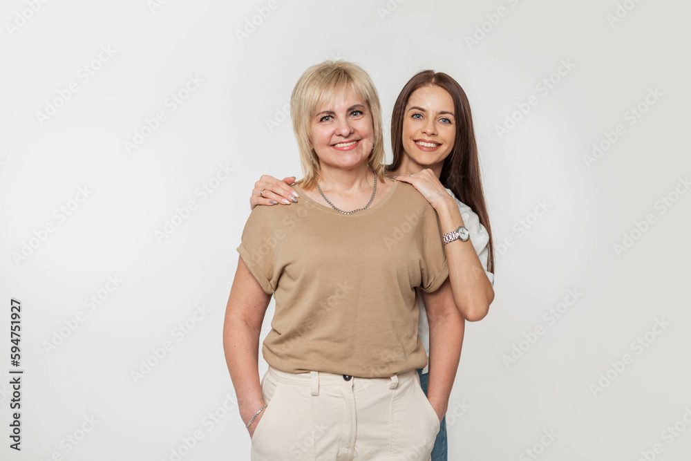 Happy woman daughter with a smile hugs a beautiful elderly mother in denim casual clothes with beige t-shirt on a white background in the studio. Happy woman daughter with a smile hugs a beautiful elderly mother in denim casual clothes with beige t-shirt on a white background in the studio.