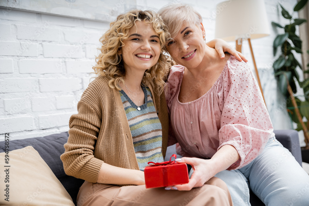 blonde woman with wavy hair embracing happy mother holding gift box while sitting on sofa at home.