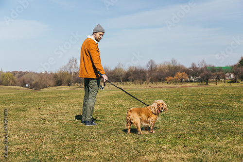 a man in a hat stands with a spaniel dog with his back into the distance along the grass in a park in autumn or spring