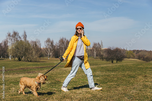 a girl in a yellow jacket and a red hat with glasses walks with a spaniel dog in the park and speaks on the phone