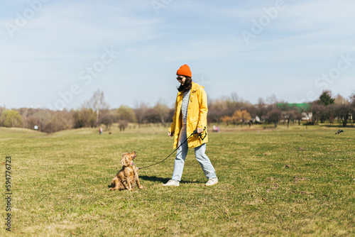 a girl in a yellow raincoat, a red hat and glasses walks with a spaniel dog in a park on the grass in sunny weather