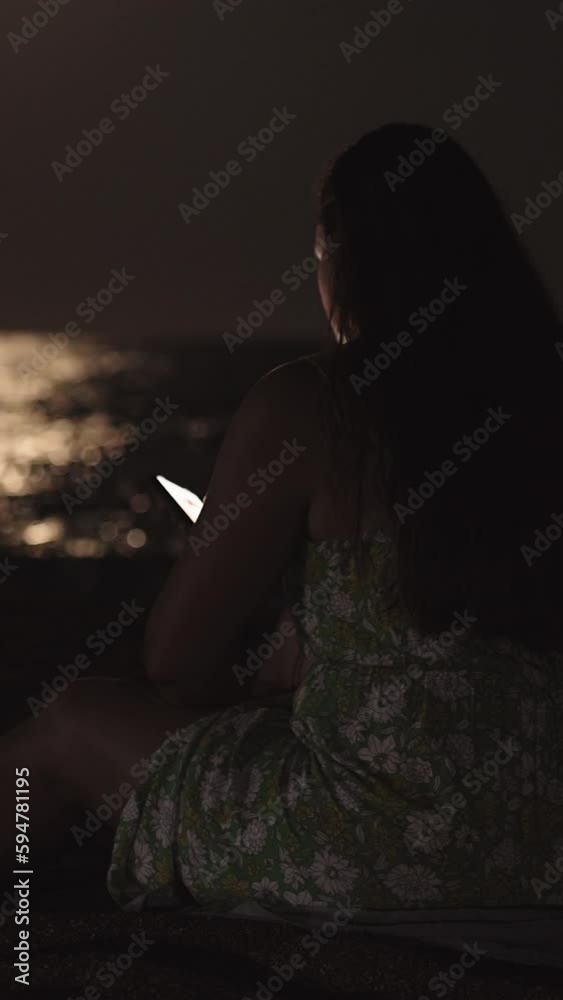 A young woman in a dress sits by the sea on the beach and looks into a smartphone. Vertical, Social Media A young woman in a dress sits by the sea on the beach and looks into a smartphone. Vertical, Social Media