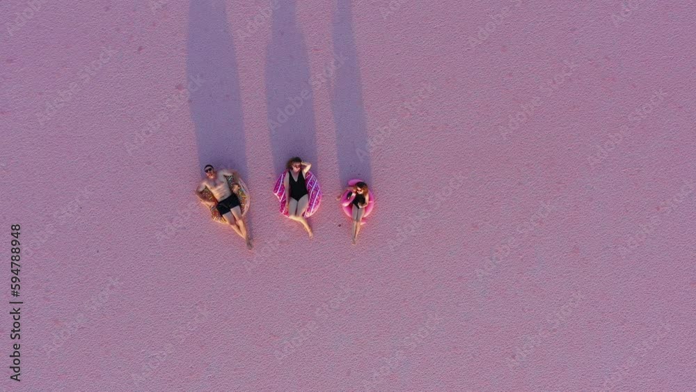 Happy summer holiday. Aerial view of young family, mom, dad, and child daughter in swimwear lying on inflatable flamingo and donuts on natural pink salt lake background and look up at rolling camera.