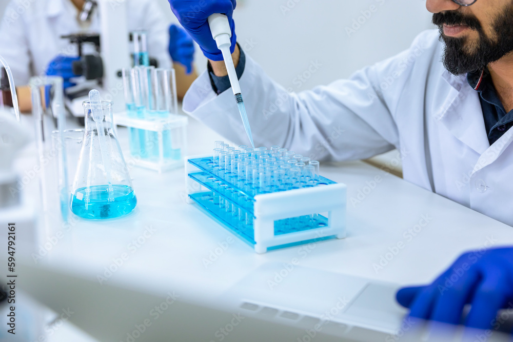 Fototapeta premium Scientist holds glass-dropper with reagents makes notes in laptop, experimenting with medical sample