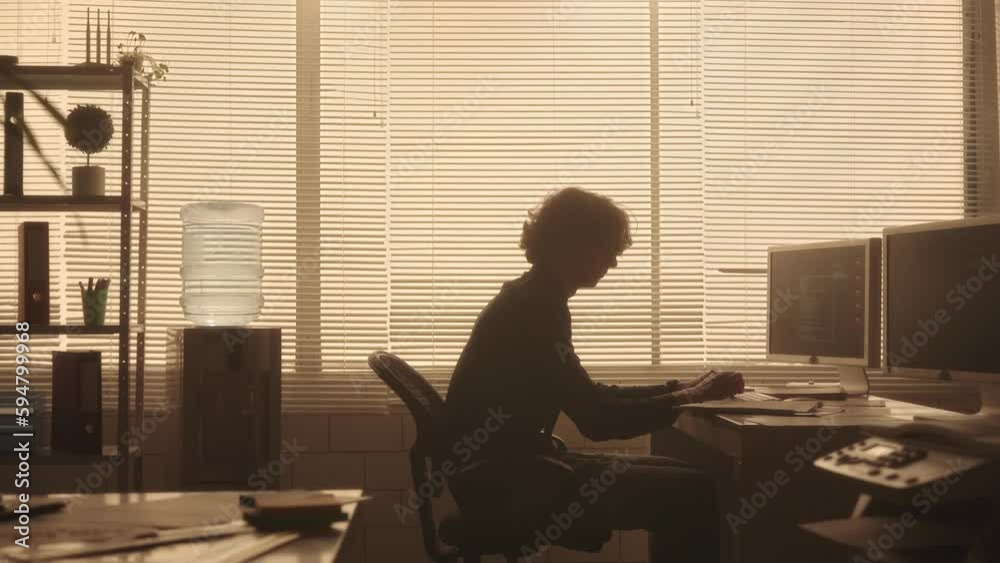 The silhouette of a young man is typing on a computer keyboard, looking through a report and frustrated scattering the sheets. The manager works sitting at a table near a window with backlight.