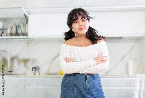 Portrait of smiling young housewife in white blouse and denim skirt standing in kitchen  posing on camera