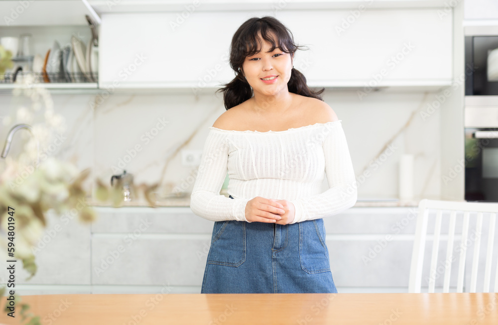 Portrait of smiling young housewife in white blouse and denim skirt standing in kitchen, posing on camera