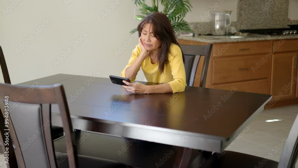 Wide shot attractive woman relaxing watching movie on mobile cell phone sitting at dark wood dining table Wide shot attractive woman relaxing watching movie on mobile cell phone sitting at dark wood dining table