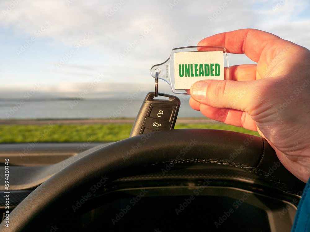 Obraz premium Driver holding car key with unleaded badge. Beautiful nature scene with ocean and mountain out of focus in the background. Travel or trip in rented car concept.