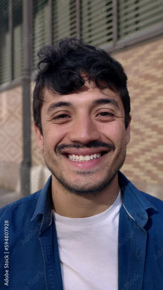 Vertical portrait of young adult latin american man smiling at camera standing outdoors. Happy hipster millennial guy against brick wall in Barcelona. People concept. Vertical portrait of young adult latin american man smiling at camera standing outdoors. Happy hipster millennial guy against brick wall in Barcelona. People concept.