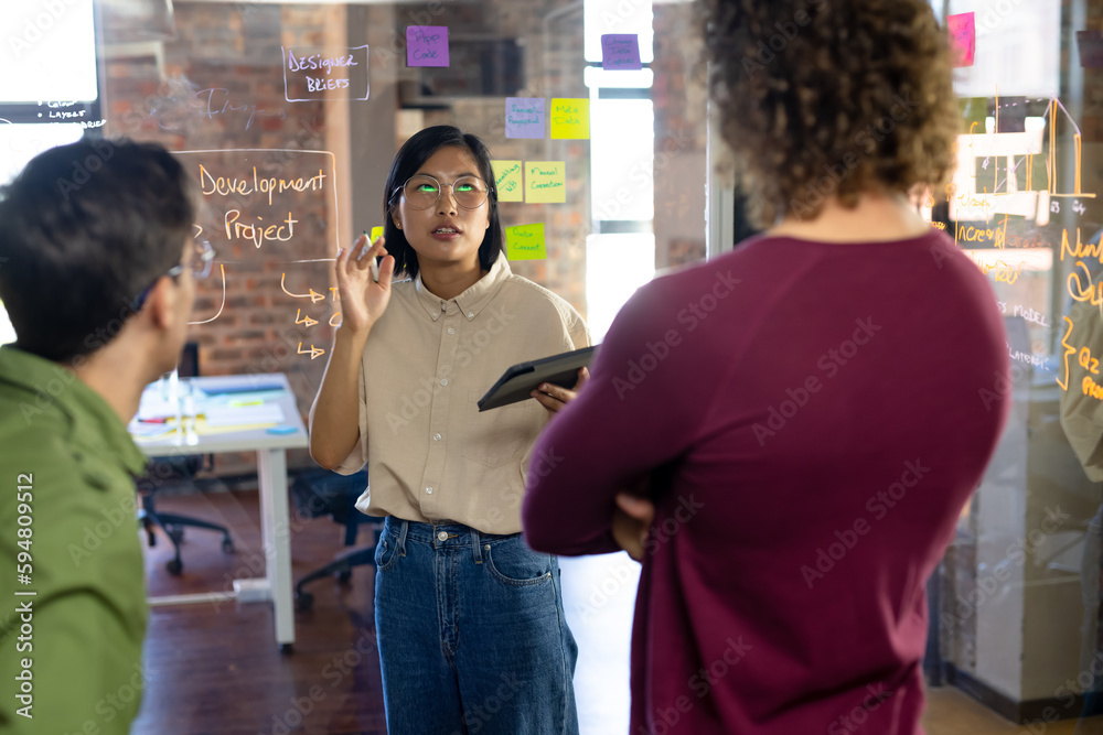Busy diverse creative colleagues using tablet, brainstorming and making notes on glass wall Busy diverse creative colleagues using tablet, brainstorming and making notes on glass wall