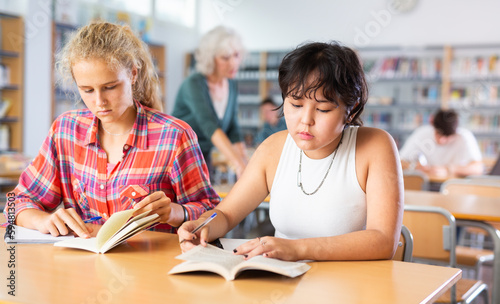 Two fifteen-year-old schoolgirls reading books and taking abstract in copybooks are doing homework in the school library © JackF