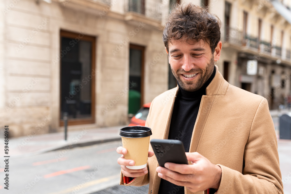 Young handsome caucasian man using smartphone smiling with a cup of coffee in his hand on the street. take away concept