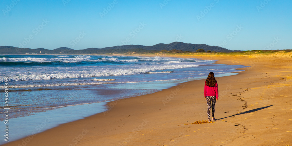 Obraz premium beautiful long haired girl walking on wooli beach during sunrise; long beach with large waves on the coast of pacific in yuraygir national park, new south wales, australia