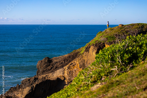 girl in white shirt and shorts standing on the grassy hill gazing at the sea in hat head national park in new south wales, australia  korogoro walking track © Jakub