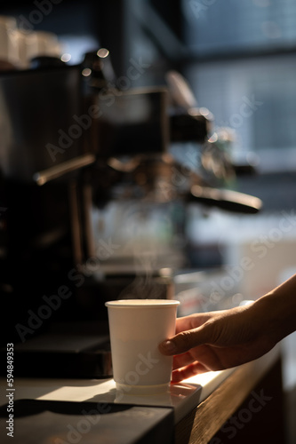 A cup of coffee sits on a table with a coffee machine in the background. © pariwatpannium